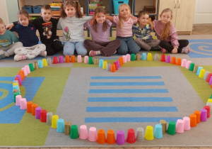 Grupka dzieci siedzi na dywanie, a przed nimi leży ułożone z kolorowych kubków serce według podanego rytmu.