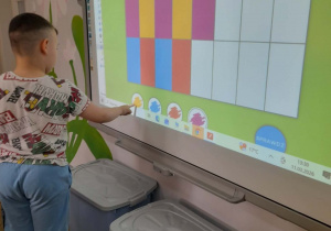 Kuba stoi przed tablicą multimedialną i kończy kolorować dywanik według podanego kodu.