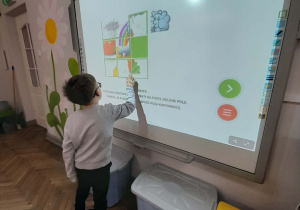 Chłopiec stoi przed tablicą multimedialną i układa obrazek przesuwając poszczególne elementy na puste zielone pole.