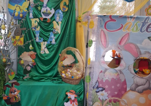 Dekoracja Wielkanocna: na tablicy okrytej zielonym materiałem przypięty jest napis Wielkanoc oraz wielkanocne symbole. Obok z jednej strony stoi wazon z baziami i pisankami z drugiej fotobudka wielkanocna. Nad dekoracją wiszą kolorowe balony w kształcie pisanek.