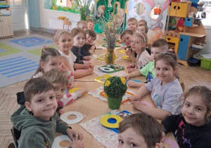 Dzieci siedzą przy wspólnym stole na którym znajdują się dekoracje wielkanocne, wieńce, jajka ze sklejki, mazaki, cekiny. W tle dekoracja wielkanocna.