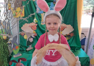 Nikola w stroju zajączka stoi na tle dekoracji z koszykiem w którym znajdują się wielkanocne gniotki.