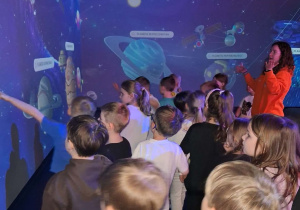 Dzieci stoją z Panią animator w strefie Stacja Galaxy Fantazjuj i oglądają interaktywne planety zasad.