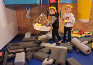 Hubert i Adaś bawią się na placu budowy. Chłopcy mają kaski na głowie.