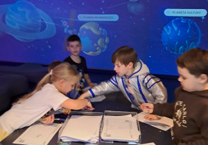 Czworo dzieci koloruje swoje kosmiczne kolorowanki. W tle interaktywna ściana, na której znajdują się planety zasad.