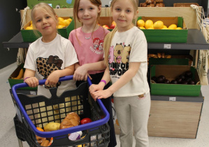 Trzy dziewczynki trzymają koszyk z zakupami. W tle stoisko z warzywami.
