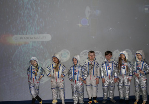 Grupka dzieci w kosmicznych skafandrach stoi obok siebie na tle interaktywnej ściany.