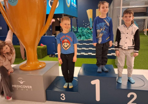 Trzech chłopców stoi na podium obok którego stoi ogromny puchar.