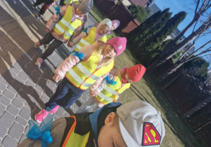 Dzieci w odblaskowych kamizelkach idą ustawione parami.