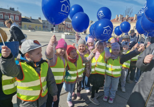 Przedszkolaki stoją na Placu Wolności i trzymają w górze niebieskie balony.