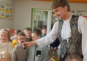 Mikołaj śpiewa do mikrofonu piosenkę Bachmana. Obok chłopca stoi Pan i grupka rówieśników.