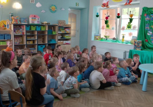 Dzieci siedzą na dywanie, a przed nimi stoi Pani z biblioteki i zaprasza przedszkolaków na spotkanie z okazji Światowego Dnia Książki i Praw Autorskich. W tle tablica z napisem Dzień Książki, okno, szafki z układankami.