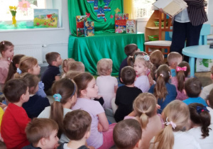 Dzieci siedzą na dywanie, a przed nimi stoi Pani z biblioteki i omawia budowę książki.