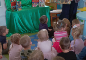 Pani stoi przed dziećmi i prezentuje książkę przyrodniczo-ekologiczną. W tle okno, tablica z napisem Dzień Książki, szafka przykryta zielonym materiałem, na której stoją książki z bajkami.