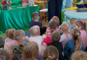 Dzieci siedzą na dywanie, a przed nimi stoi Pani z biblioteki, która trzyma książkę o emocjach pt. "Kolorowy potwór".