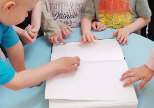Grupka dzieci z grupy IV ogląda przy stoliku książkę dla osób niewidomych.