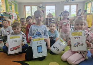 Dzieci siedzą na dywanie. Olek, Ala, Szymon i Lila trzymają plansze z numerami alarmowymi.