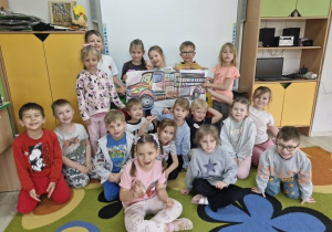 Przedszkolaki stoją przed tablicą multimedialną trzymając w dłoniach dużą kolorowankę przedstawiającą wóz strażacki.