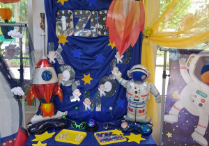 Dekoracja z okazji Dnia Kosmosu: na tablicy okrytej granatowym materiałem przypięty jest napis Dzień Kosmosu i żółte gwiazdki. Na szafce stoi balon w kształcie kosmonauty i rakiety. Nad tablicą wiszą lampiony w kształcie planet.