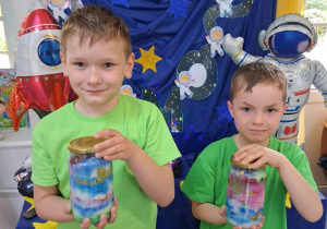 Marcel i Bartek stoją na tle dekoracji i trzymają słoiki wypełnione kolorową watą i brokatem.
