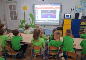 Dzieci siedzą przy stolikach i oglądają prezentację na tablicy multimedialnej "W kosmosie".
