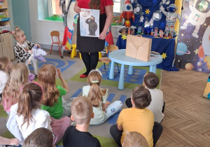 Dzieci siedzą na dywanie, przed nimi stoi pani i trzyma zdjęcie Misia Wojtka. Obok na stoliku stoi Teatrzyk Kamishibai.