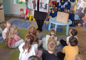 Przedszkolaki siedzą na dywanie i słuchają pani, która opowiada o Misiu Wojtku i generale W. Andersie. Pani trzyma zdjęcia Misia Wojtka i generała W. Andersa