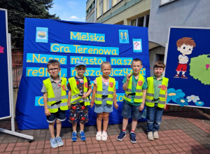 Pięcioro dzieci stoi na tle dekoracji z napisem Miejska Gra Terenowa "Nasze miasto, nasz region, nasza ojczyzna”.