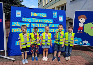 "Słoneczka" stoją na tle dekoracji z napisem Miejska Gra Terenowa "Nasze miasto, nasz region, nasza ojczyzna”.