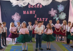 Na środku sceny troje dzieci stoi obok siebie i gra na tamburynach, a pozostałe przedszkolaki stoją w półkolu. W tle dekoracja.