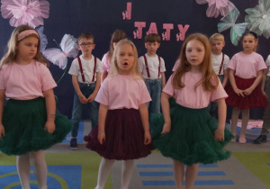 Trzy dziewczynki śpiewają piosenkę na środku sceny. Za nimi stoją rówieśnicy w półkolu.