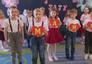 Grupka dzieci recytuje wiersz na środku sceny. Czworo przedszkolaków trzyma serca tworząc napis - Mamo. W tle inne przedszkolaki stoją na tle dekoracji.