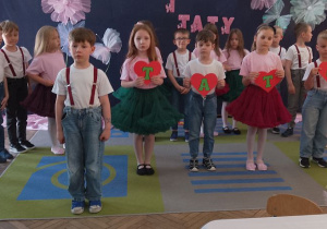 Chłopiec na środku sceny mówi fragment wiersza, a obok stoi czworo rówieśników z sercami tworzącymi napis - Tato. W tle inne przedszkolaki stoją na tle dekoracji.