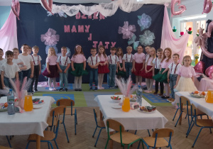 Dzieci stoją w półkolu na tle dekoracji. Przed dziećmi stoją trzy stoliki z poczęstunkiem.