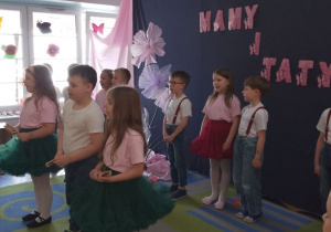 Dzieci śpiewają piosenkę. Troje przedszkolaków stoi na środku sceny trzymając instrumenty. W tle fragment dekoracji, okno.