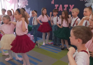 Trzy dziewczynki stoją na środku sceny, a za nimi rówieśnicy. Dzieci śpiewają piosenką i naśladują ruchem jej treść.