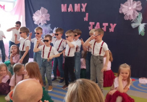 Chłopcy tańczą w ciemnych okularach na środku sceny, a obok przykucnęły dziewczynki.