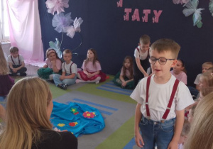 Mikołaj w roli narratora recytuje fragment wiersza "Żuraw i Czapla", Tosia w roli Czapli siedzi przy toaletce. Pozostałe dzieci siedzą na dywanie. W tle dekoracja.