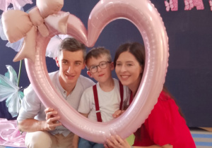Mikołaj razem z rodzicami pozuje do zdjęcia trzymając balonowe serce. W tle dekoracja.