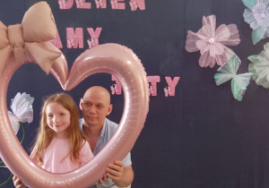Emma razem a tatą pozuje do zdjęcia trzymając balonowe serce. W tle dekoracja.