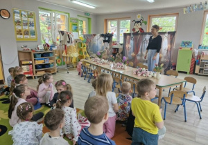 Dzieci siedzą na dywanie. Pani stoi przed stołem pełnym kwiatów i opowiada przedszkolakom na czym będą polegały dzisiejsze zajęcia.