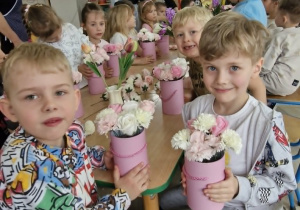Lucjan i Olek siedząc przy stoliku prezentują wykonane przez siebie bukiety kwiatów.