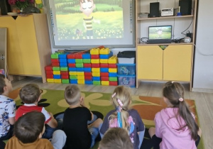 Dzieci siedząc na dywanie oglądają film edukacyjny.