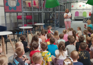 Dzieci siedzą na podłodze, a Pani stoi przed nimi z mikrofonem w ręku i przypomina zasady bezpiecznej zabawy.