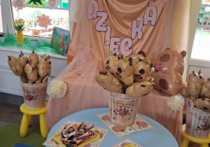 Dekoracja z okazji Dnia Dziecka: na tablicy okrytej beżowym materiałem przypięty jest napis Dzień Dziecka z motywem kapibary, białe bibułowe kwiaty oraz balon w kształcie kapibary. Przed tablicą stoi niebieski stolik na którym znajdują się upominki ufundowane przez rodziców: zegarki z motywem kapibary, papierowe kapibarowe okulary i balony w kształcie kapibary.