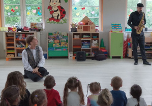 Dzieci siedzą na dywanie i słuchają utworu granego przez muzyka na saksofonie. Obok siedzi Pan prowadzący zajęcia.