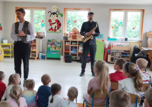 Dzieci siedzą na dywanie, a przed nimi stoi Pan prowadzący audycję muzyczną. Obok stoi muzyk trzymajacy saksofon.