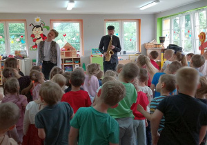 Dzieci na stojąco poruszają się przy dźwiękach saksofonu. Przed nimi gra muzyk, a obok stoi Pan prowadzący audycję.