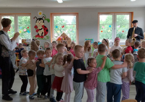 Muzyk gra na saksofonie, a dzieci razem z Panem prowadzącym audycję utworzyły pociąg i poruszają się przy dźwiękach muzyki.