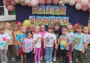 Pszczółki pozują do grupowego zdjęcia na tle napisu zabawy sportowe i baneru ozdobionego balonami.